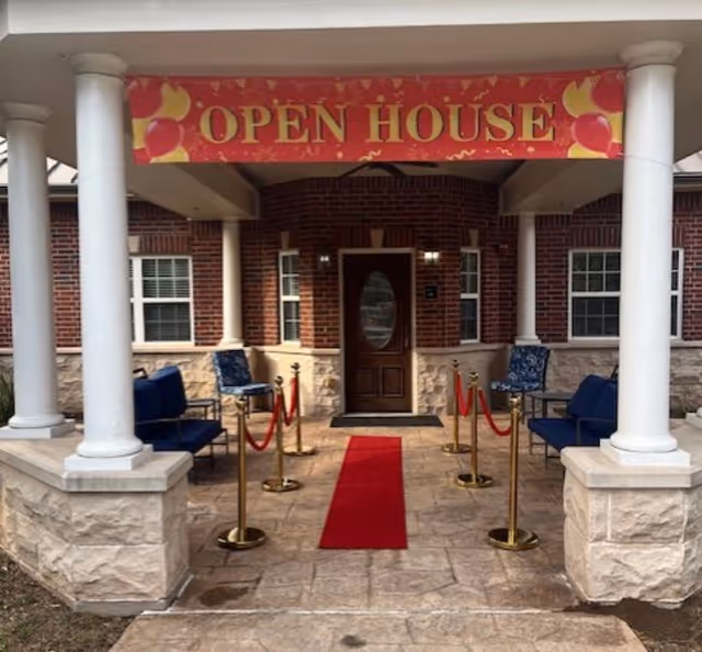Front entrance of a brick building with white columns, a red carpet leading to a wooden door and an 'OPEN HOUSE' banner overhead.