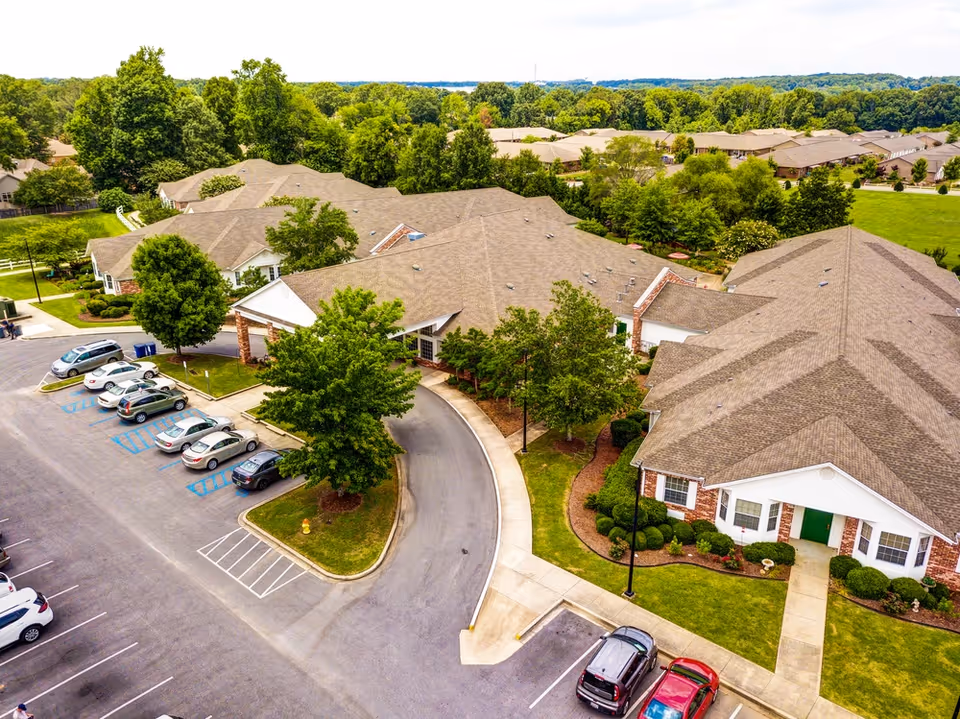 Aerial view of The Renaissance of Florence senior living facility showing multiple connected buildings with brown roofs surrounded by green trees and landscaped lawns. Several cars are parked in the parking lot adjacent to the buildings.