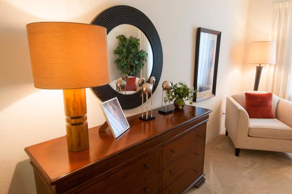 A cozy living room corner featuring a wooden dresser with a large round black-framed mirror above it. On the dresser are a table lamp with a beige shade, a framed photo, decorative metallic sculptures, and a small potted plant. To the right, there is a beige armchair with a red cushion and a floor lamp with a beige shade next to a window with light curtains.