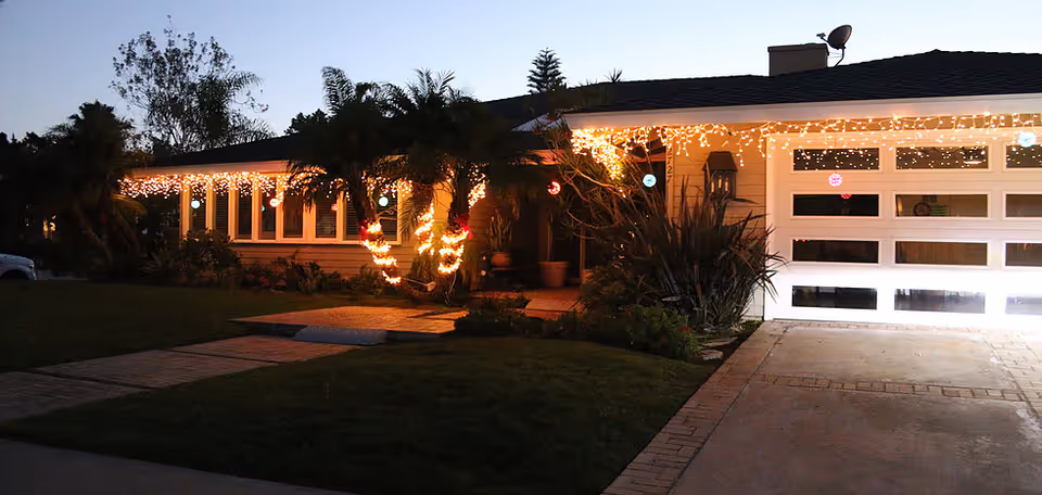 A single-story house decorated with string lights along the roofline and wrapped around palm trees in the front yard during dusk.