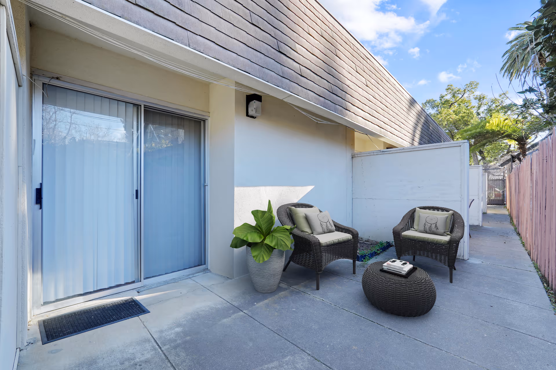 Outdoor patio area with two wicker chairs with cushions and a small wicker table with books on it, next to a sliding glass door with vertical blinds. There is a large potted plant beside the chairs and a wooden fence along the right side of the concrete pathway.