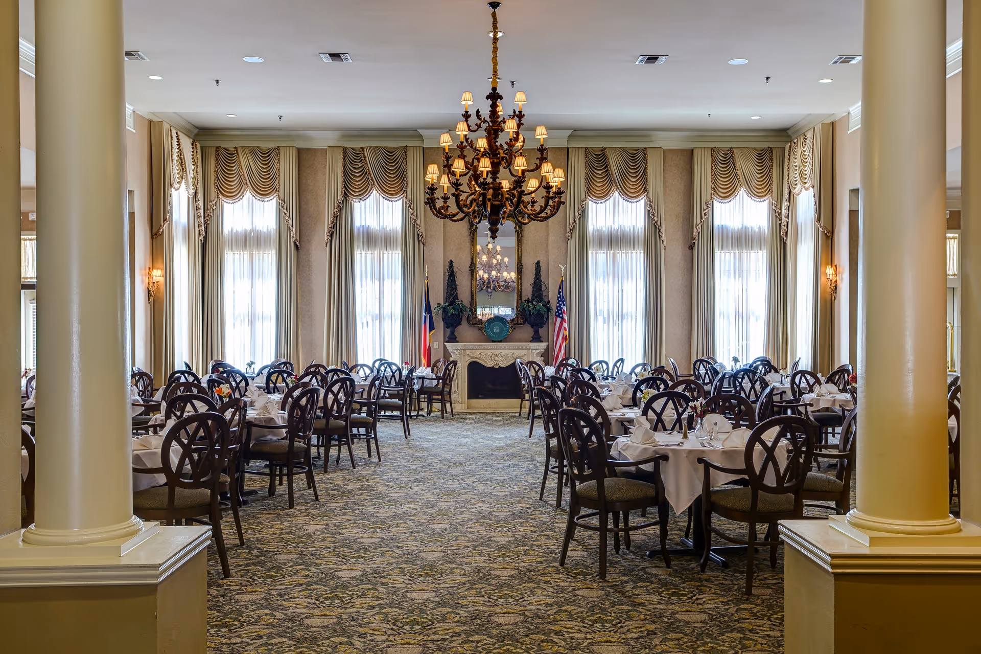 Elegant dining room with multiple round tables covered with white tablecloths and set with napkins and glassware. The room features tall windows with draped curtains, a large ornate chandelier hanging from the ceiling, two large columns in the foreground, and a fireplace with decorative items and flags on either side.