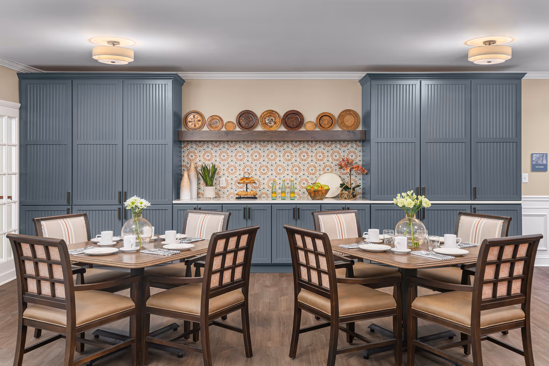 A dining area with two wooden tables, each surrounded by four chairs with cushioned seats and backs. The tables are set with white plates, cups, and silverware, and each has a glass vase with white flowers. Behind the tables is a wall with blue-gray cabinets and a decorative backsplash featuring a patterned tile design. Various decorative plates and vases are displayed on a shelf above the countertop.