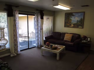 A cozy living room with a dark brown leather sofa adorned with a striped pillow, a white coffee table with decorative items, and a large painting of a landscape on the wall. The room has sliding glass doors with patterned curtains leading to an outdoor patio with a table and chairs. There is a window with blinds and a birdcage on a stand near the door.