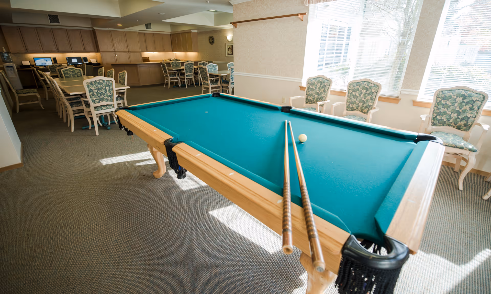 Interior common room with a pool table and cues in the foreground, dining tables, computer stations and upholstered chairs by windows in the background.