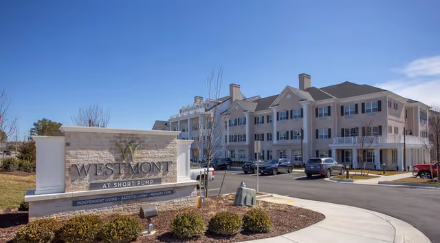 Exterior view of The Westmont at Short Pump senior living facility showing a large three-story building with multiple windows and balconies, a parking lot with several cars, and a stone sign in the foreground displaying the facility's name and services offered.