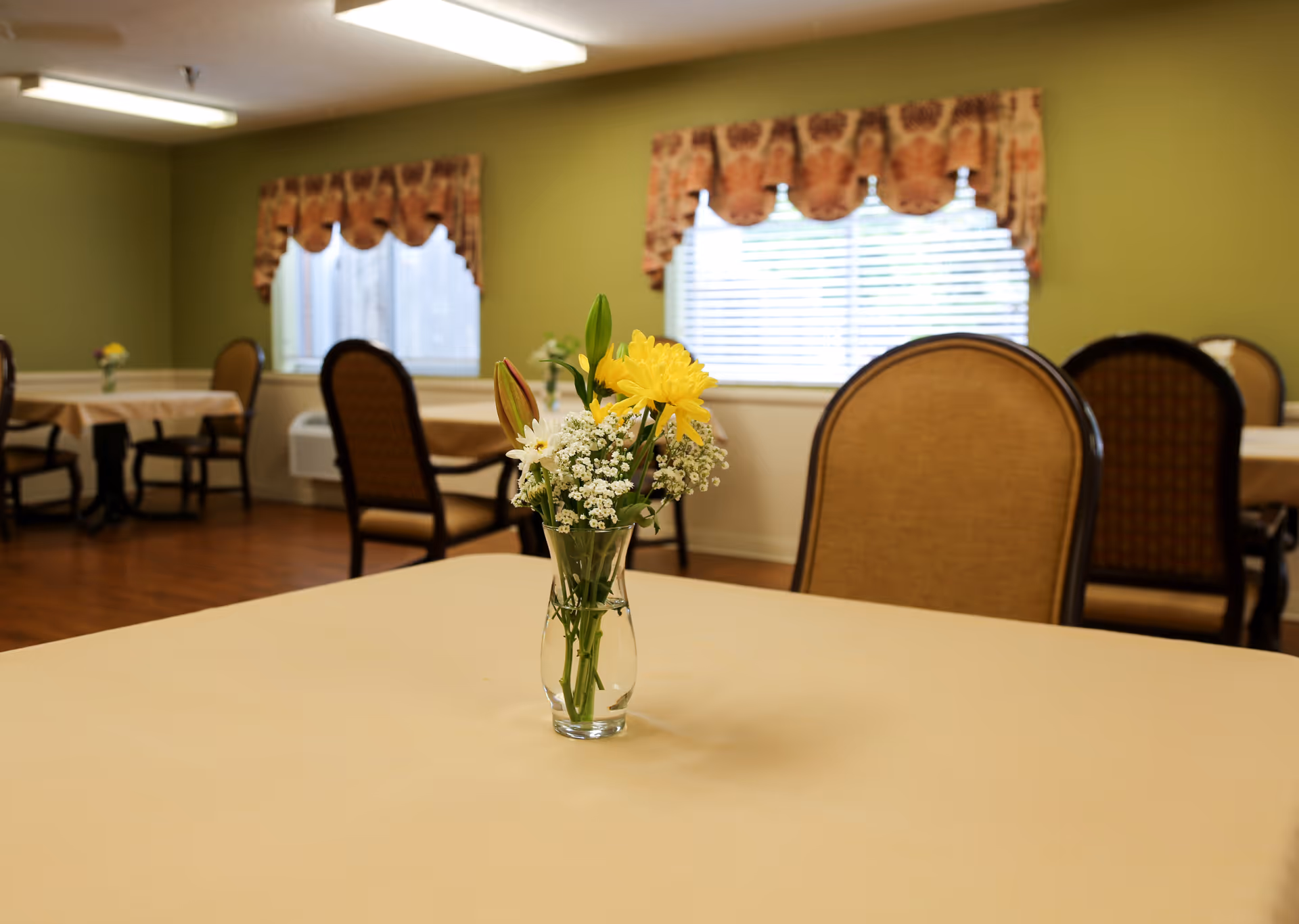 A dining room with tables covered in beige tablecloths, each table decorated with a small vase of fresh flowers. The room has green walls, wooden flooring, and windows with patterned valances allowing natural light to enter.