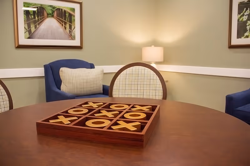 A cozy interior room with a round wooden table featuring a large tic-tac-toe game set. Surrounding the table are cushioned chairs, including a blue armchair with a plaid pillow. The walls are light green with white trim, decorated with framed nature photographs and a lit table lamp in the corner.