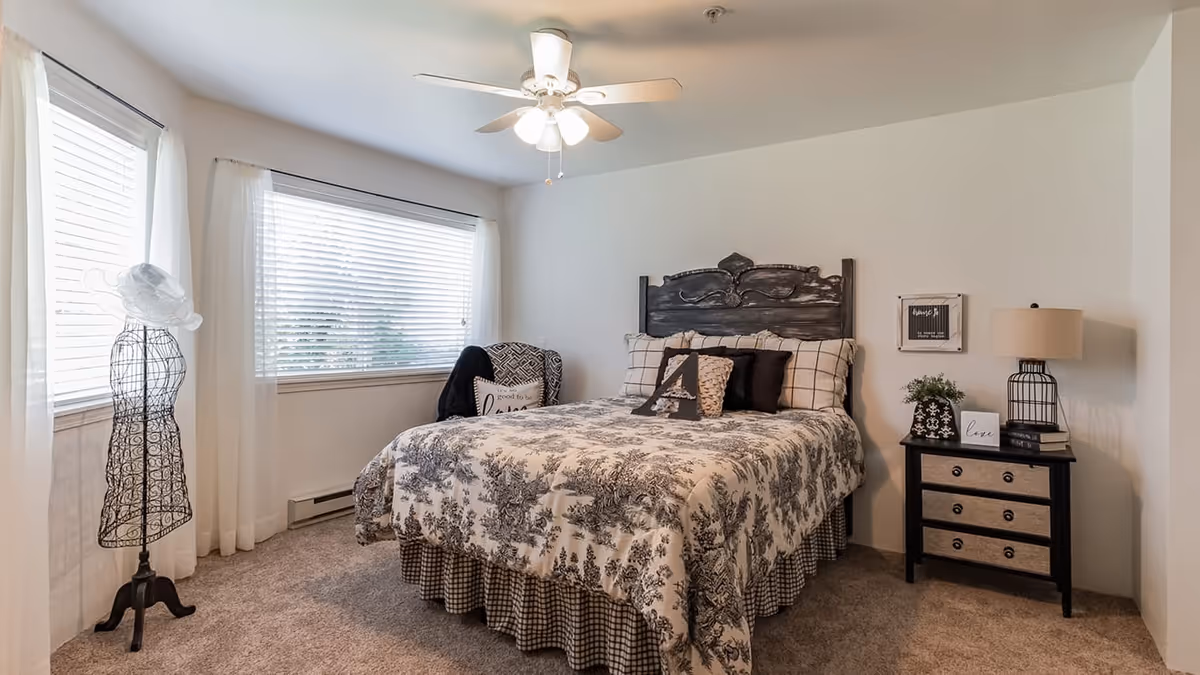 A cozy bedroom with a bed featuring a patterned comforter and multiple pillows, a decorative headboard, a nightstand with a lamp and small plants, a chair with a throw blanket and pillow, and a dress form near two windows with white curtains and blinds.