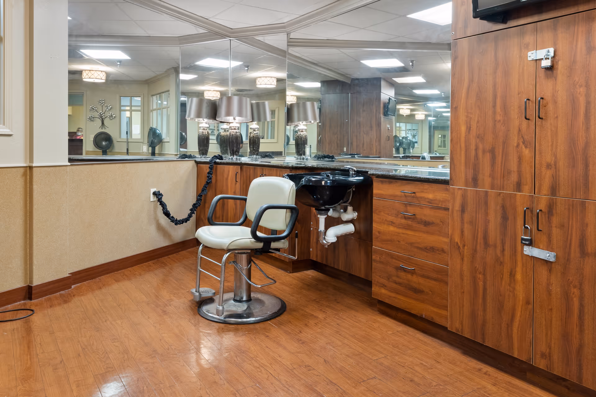 Interior view of a salon area in a senior living facility featuring a single salon chair in front of a counter with a black wash basin. The counter and cabinets are made of wood, and a large mirror covers the wall behind the counter. The floor is wooden, and the room is well-lit with ceiling lights and table lamps reflected in the mirror.