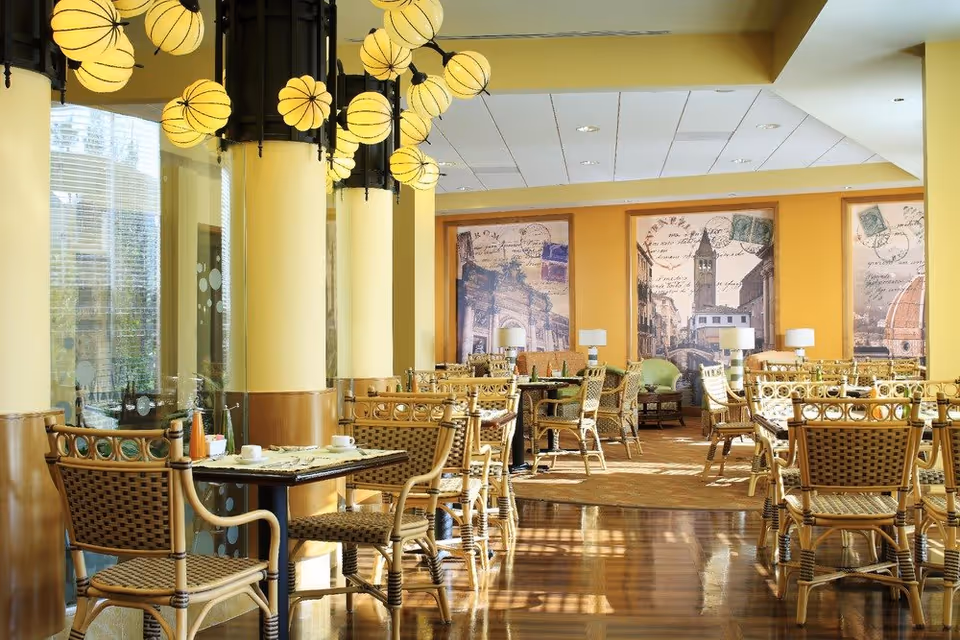 A bright dining room with multiple tables and wicker chairs arranged neatly. The room features large windows allowing natural light to flood in, yellow walls, and decorative wall art with vintage postcard designs. Unique round pendant lights hang from the ceiling, and the floor is polished wood.