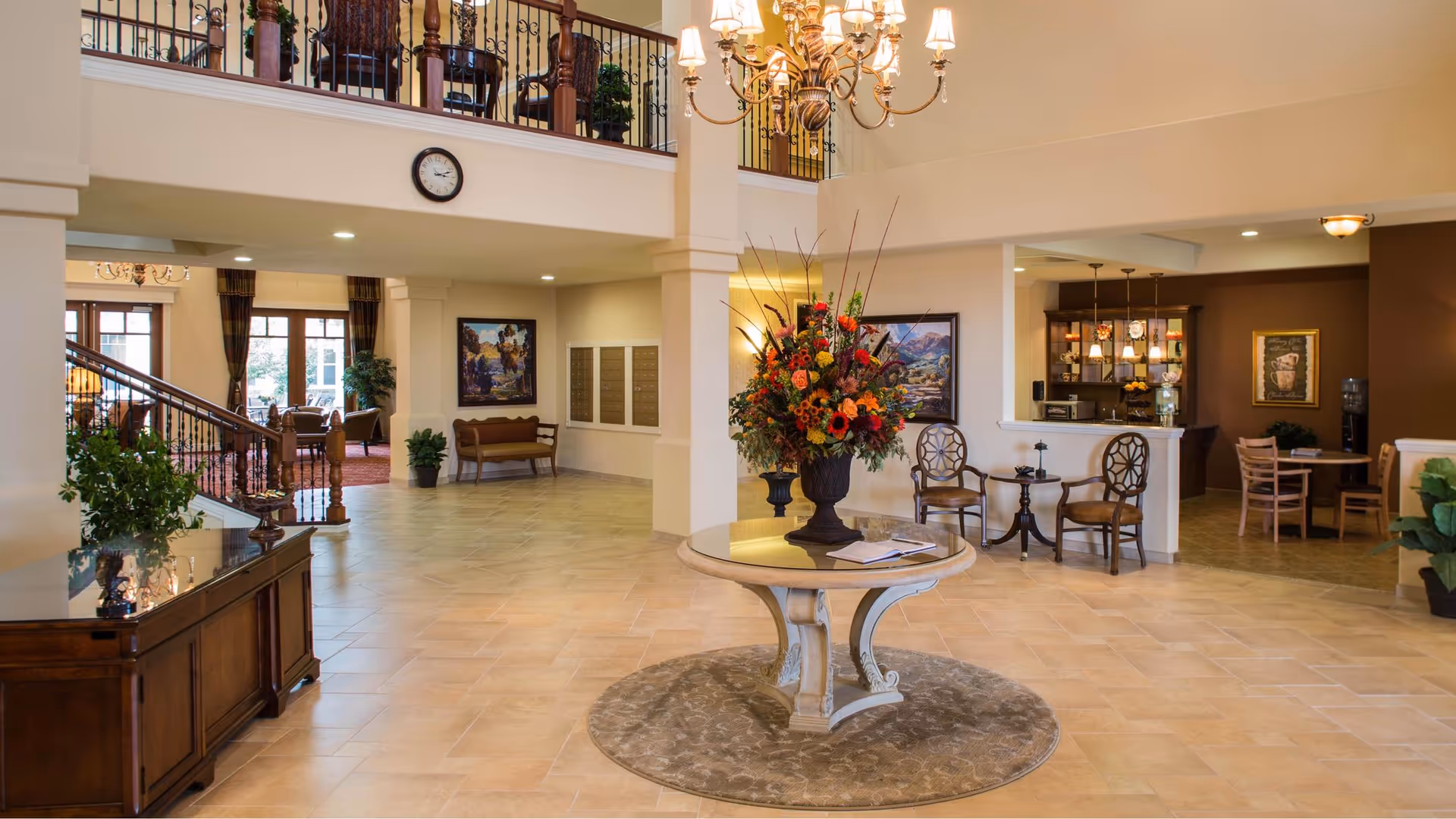 Spacious and well-lit senior living facility lobby with a round table in the center holding a large floral arrangement. The area features tiled flooring, a staircase with wooden railing, seating areas with chairs and benches, framed artwork on the walls, and a chandelier hanging from the ceiling. There is a clock on the wall above the staircase and a reception or dining area visible in the background.