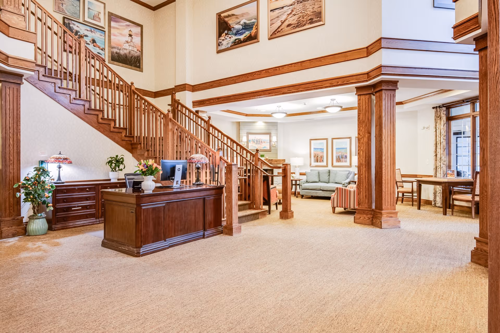Interior view of a senior living facility lobby with a wooden reception desk, staircase with wooden railing, and a seating area with a sofa, armchair, and framed artwork on the walls. The space features warm wood accents and soft lighting.