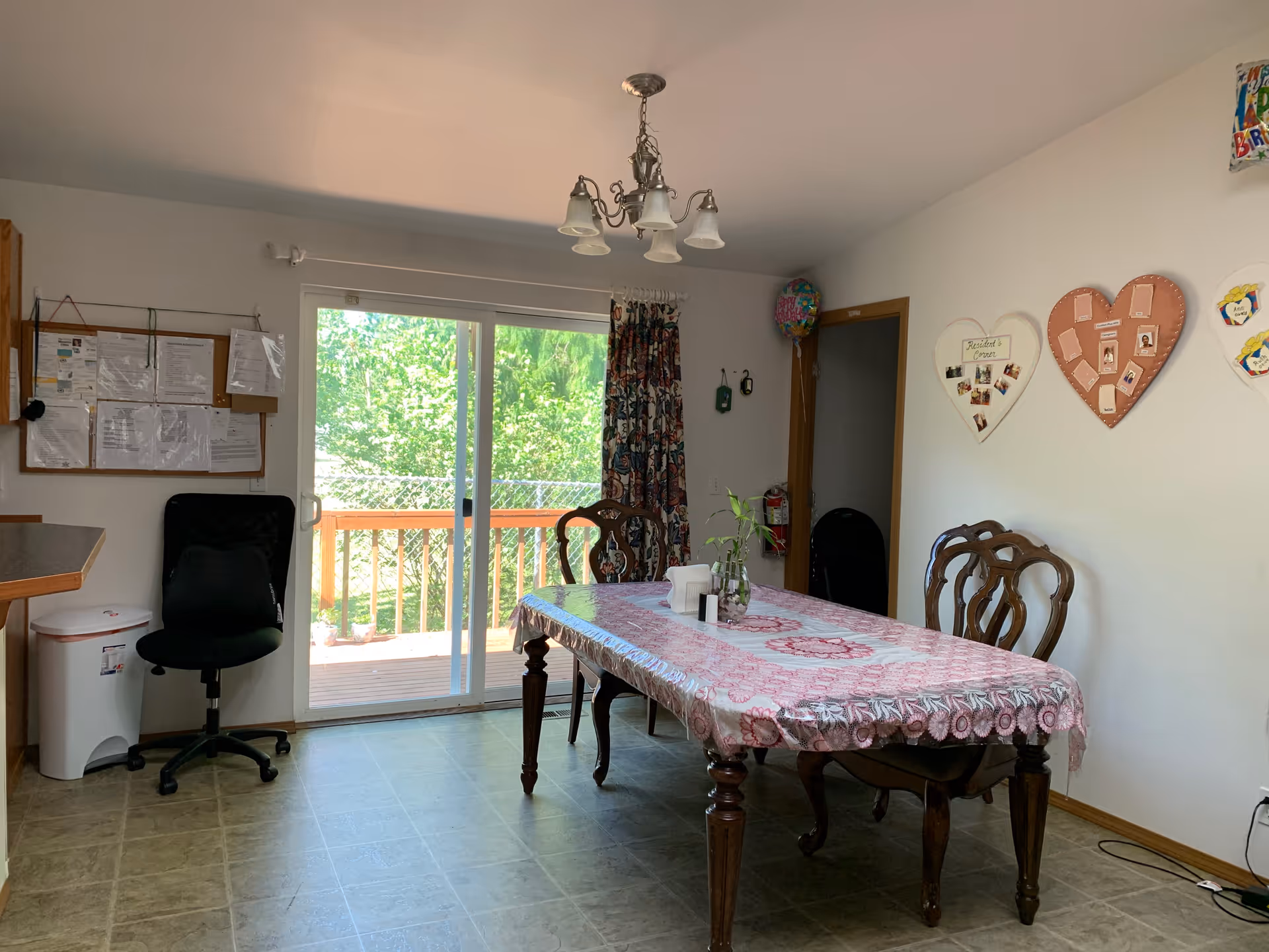 A dining area with a rectangular wooden table covered with a pink patterned tablecloth. Four wooden chairs surround the table. On the table, there is a small vase with a plant and some napkins. Behind the table, there is a sliding glass door leading to an outdoor deck with greenery visible outside. To the left, there is a black office chair and a bulletin board with papers pinned on the wall. The walls are white with some heart-shaped photo displays and decorations. A ceiling light fixture with five lamps hangs above the table.