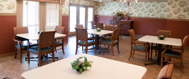 Dining room with several square tables and wooden chairs, floral wallpaper, and small flower centerpieces.