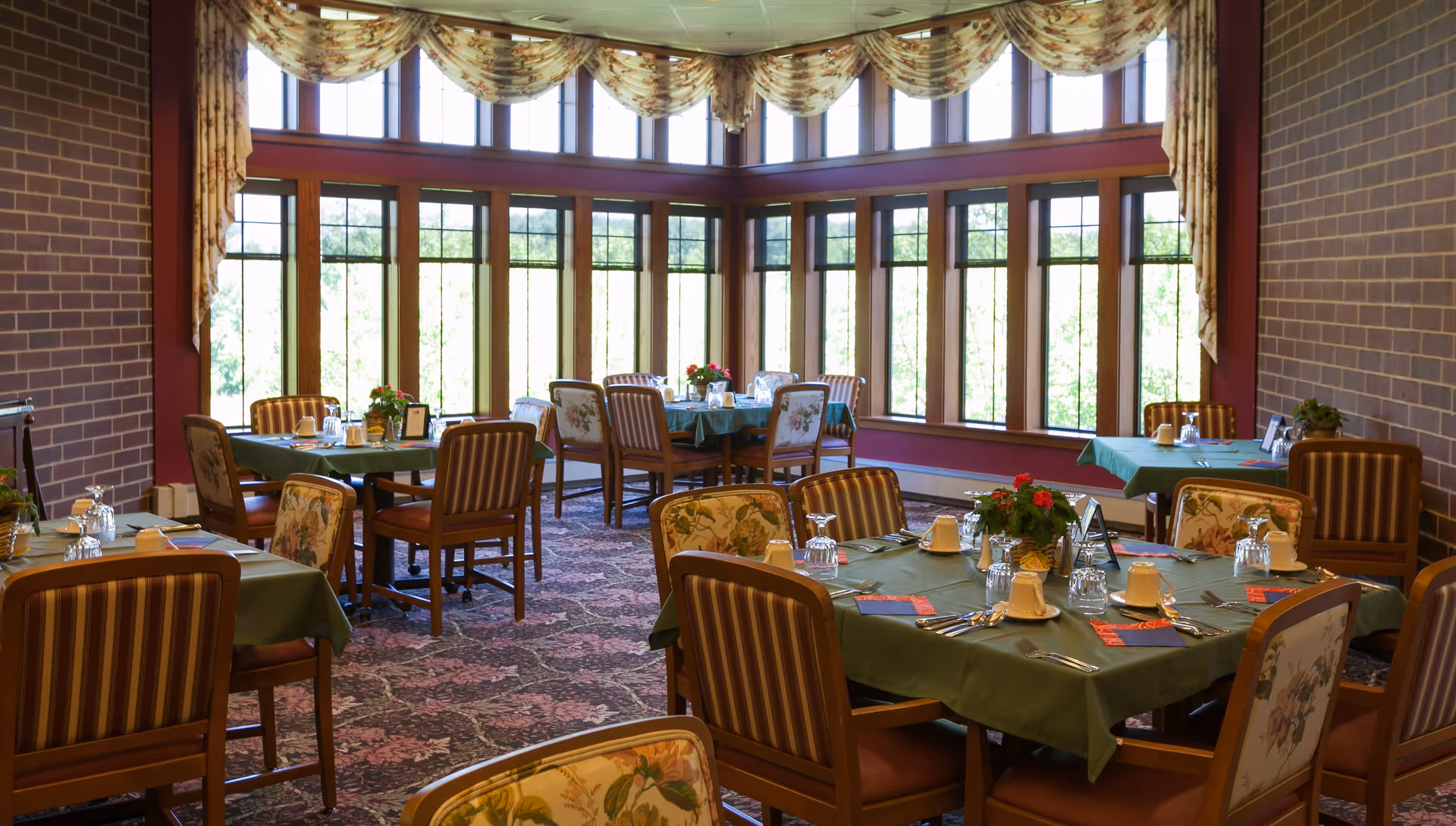 A dining room with several tables covered in green tablecloths, set with glasses, plates, and silverware. The room has large windows with floral valances, allowing natural light to fill the space. The chairs have floral and striped upholstery, and there are small flower arrangements on the tables.