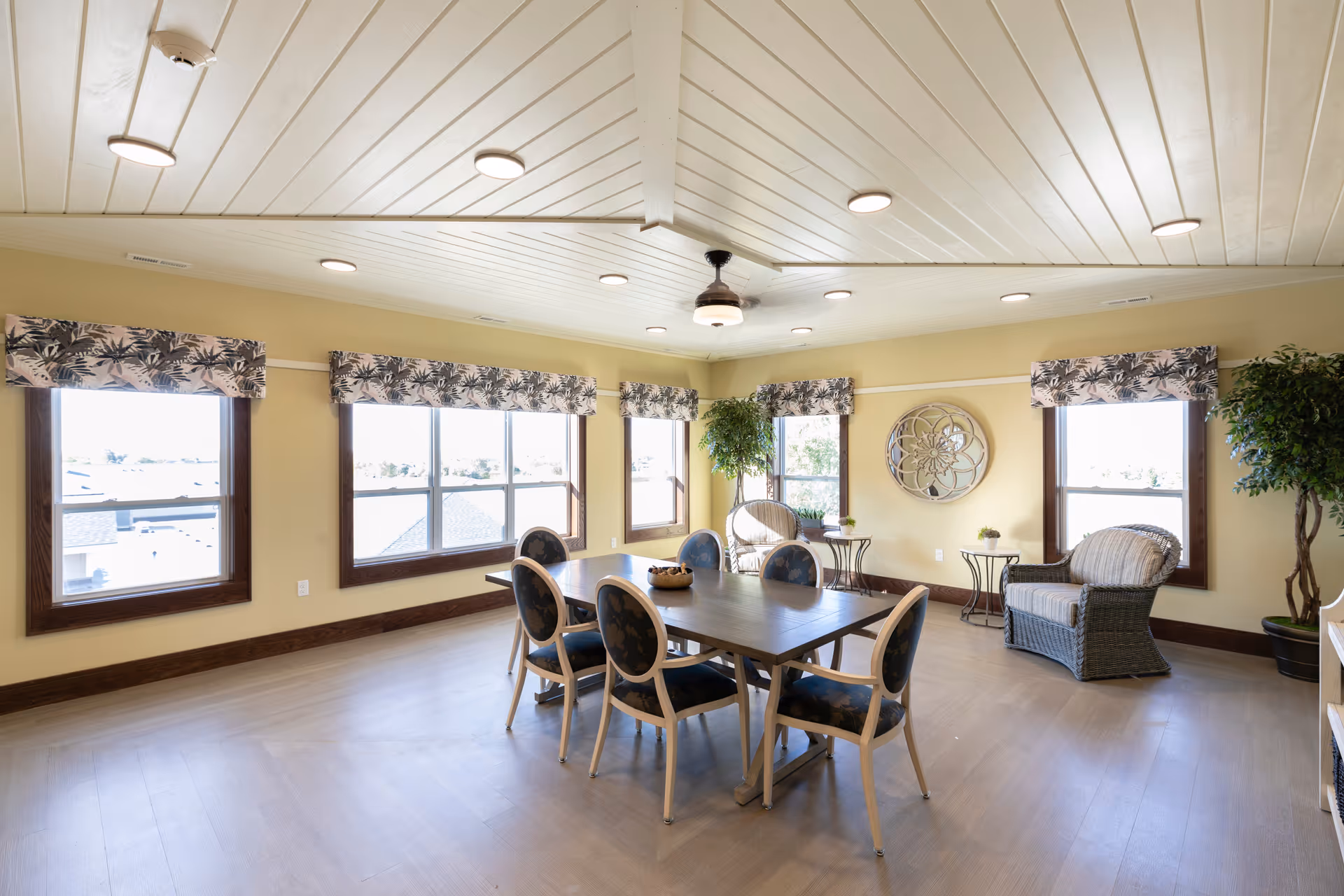 A bright and spacious common area with large windows featuring floral valances, a rectangular wooden table surrounded by six chairs with dark cushions, two wicker armchairs, small side tables, potted plants, and a decorative wall mirror. The ceiling is white with recessed lighting and a ceiling fan in the center.