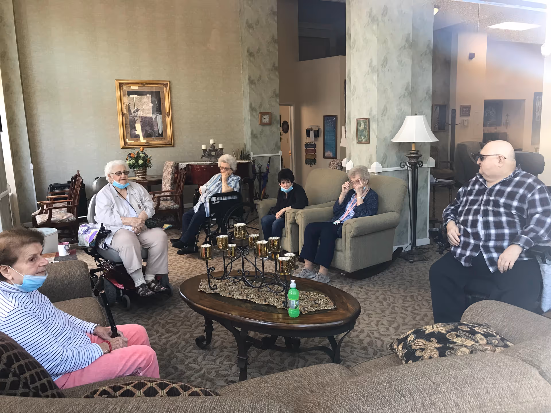 A group of elderly people sitting in a living room area of a senior living facility. The room has comfortable armchairs and sofas arranged around a wooden coffee table with decorative candle holders and a bottle of hand sanitizer. The walls are decorated with framed pictures and a lamp stands beside one of the armchairs.