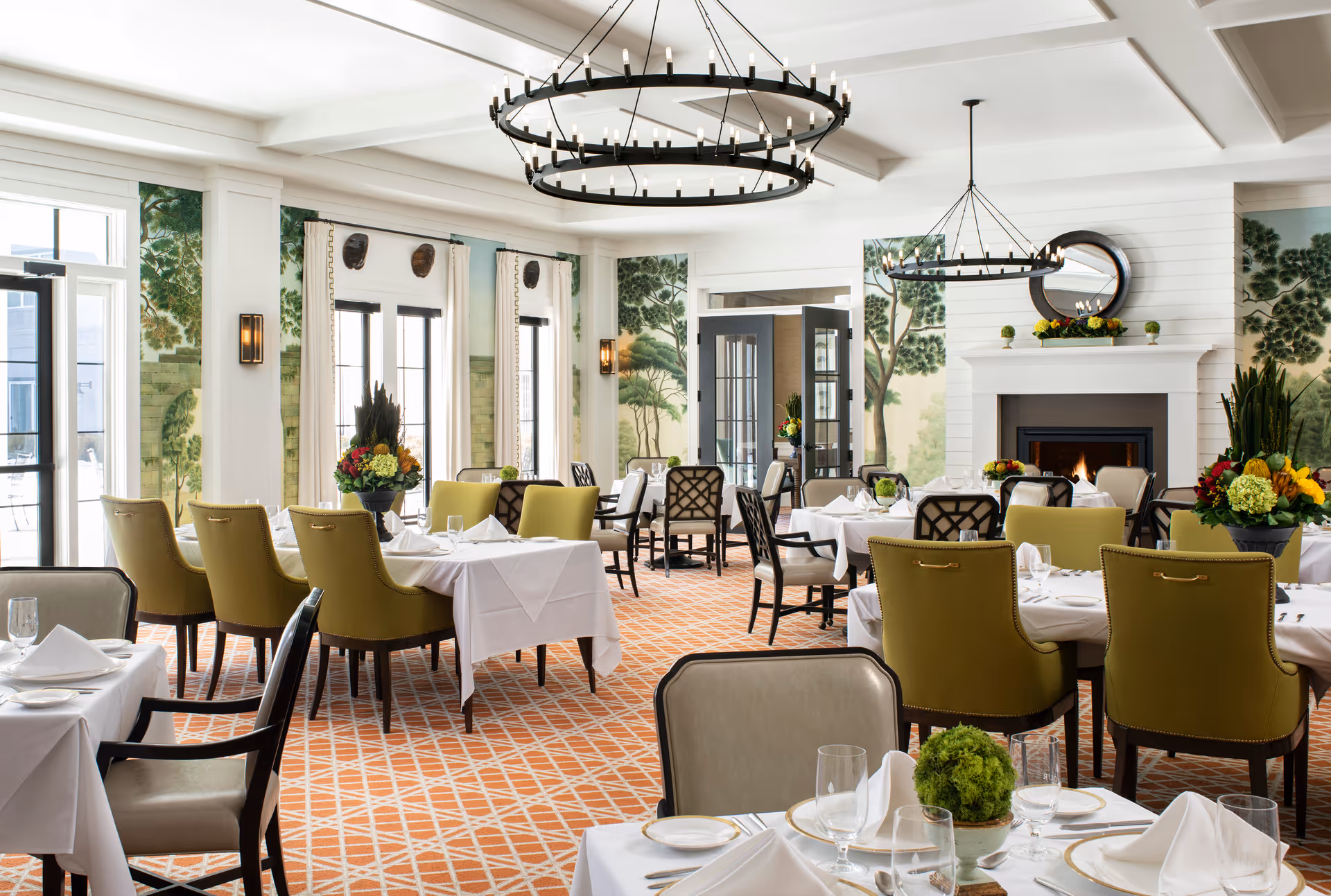 A bright and elegant dining room with tables covered in white tablecloths, set with plates, glasses, and napkins. The room features green upholstered chairs, large windows with nature-themed wall murals, two black circular chandeliers, and a fireplace with a round mirror above it. Floral centerpieces decorate the tables.