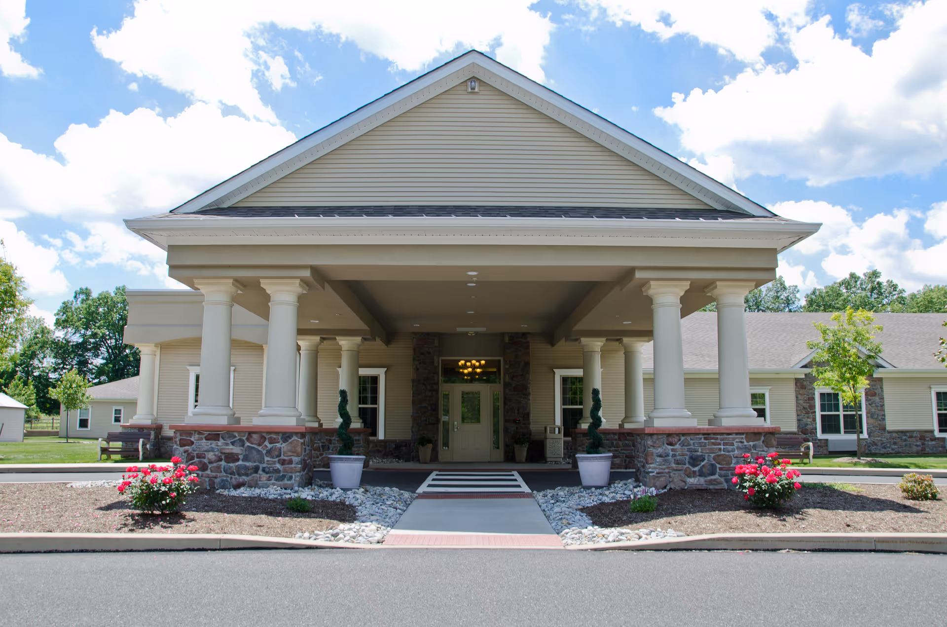 Front exterior view of a senior living facility named Hidden Meadows On the Ridge, featuring a covered entrance with white columns, stone accents, potted plants, and a clear blue sky with clouds.
