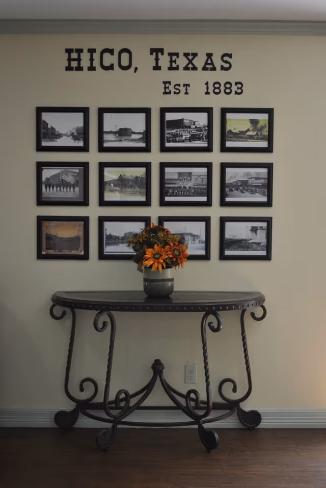 Interior wall display reading 'HICO, Texas Est 1883' over a grid of framed historic photos and a console table with a vase of sunflowers.