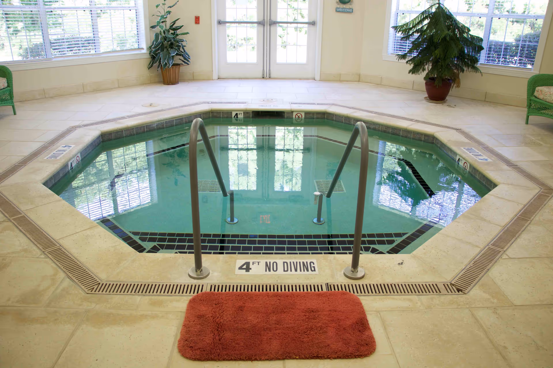 Indoor small octagonal pool or hot tub with metal handrails, surrounded by beige tiled floor. There is a red bath mat in front of the pool entrance. Large windows and glass doors allow natural light into the room, with potted plants placed near the walls.