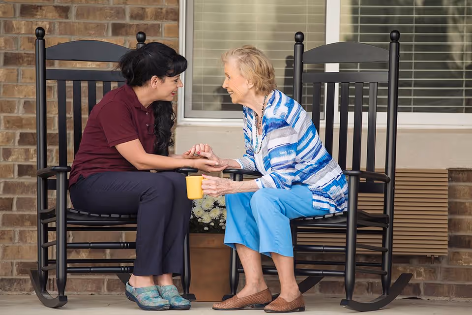 Two women sit on rocking chairs on a porch, holding hands while one holds a yellow mug.