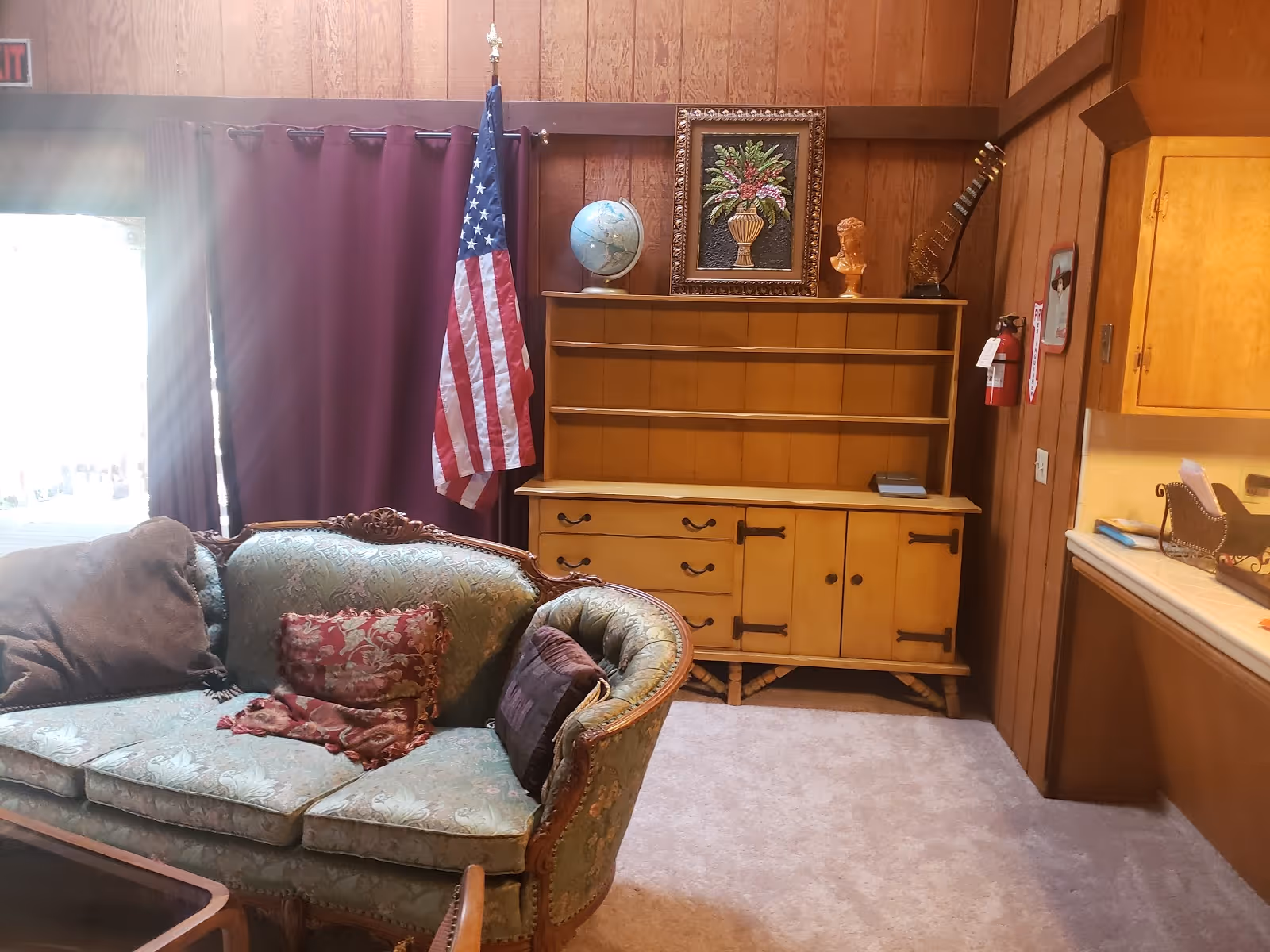 Wood-paneled living room with a vintage upholstered sofa, wooden hutch topped with a globe and framed art, and an American flag.