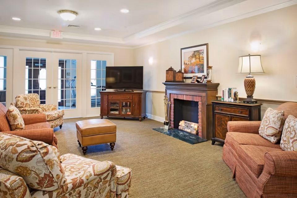 A cozy living room with patterned armchairs, a brown sofa, a leather ottoman, a wooden TV stand with a flat-screen TV, and a decorative fireplace with framed pictures and lanterns on the mantel. The room has beige walls, carpeted floor, and a table lamp on a side table next to the sofa. French doors with glass panes lead to another room.
