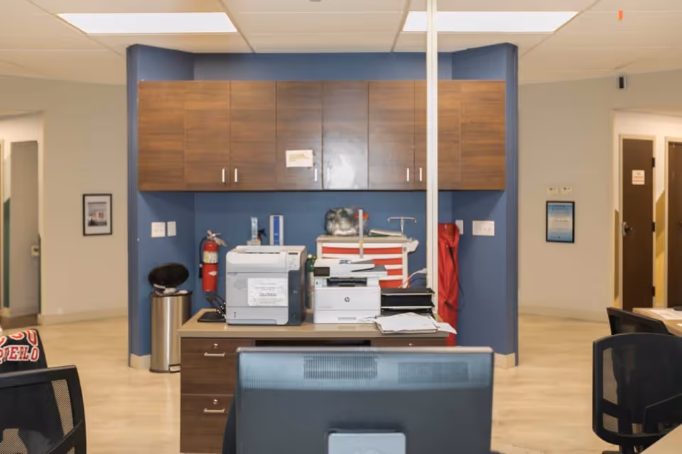 Office area with a desk holding two printers, a stack of papers, and office supplies. Behind the desk is a wall with wooden cabinets, a fire extinguisher, and a red emergency cart. The room has beige walls and a tiled floor, with doorways visible on either side.