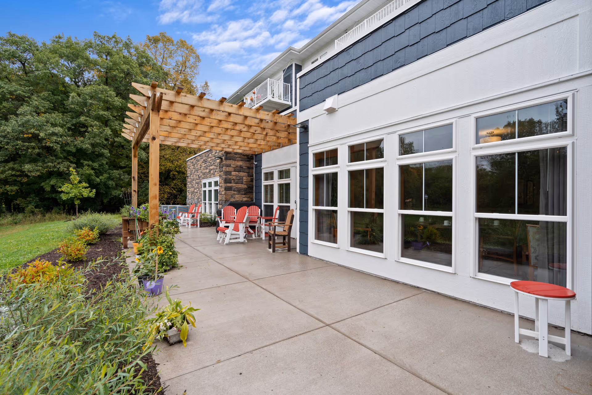 Outdoor patio area at a senior living facility with a wooden pergola, several red and white Adirondack chairs, a small white table with a red top, and a garden with various plants and flowers along the side. The building has large windows and a stone accent wall, with trees and greenery in the background under a partly cloudy sky.