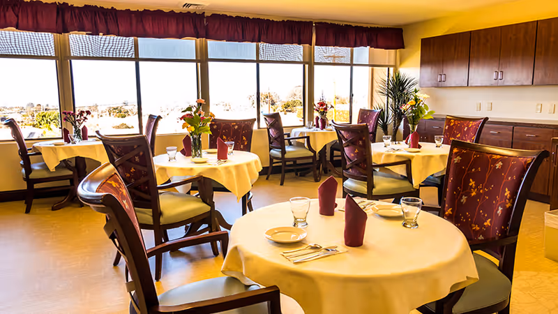 A bright dining room with round tables covered in white tablecloths, each set with plates, glasses, silverware, and folded burgundy napkins. The chairs have wooden frames with floral patterned upholstery. Large windows with a valance let in natural light and offer a view of the outdoors. There are cabinets and a countertop along one wall, and a few flower arrangements on the tables and near the windows.