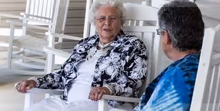 Two elderly women sitting and talking on white rocking chairs on a porch or patio area.