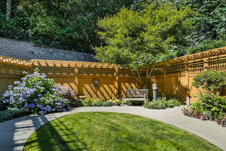 Sunlit enclosed garden courtyard with a wooden bench, flowering shrubs, a pergola-style wooden fence, curved lawn and paved walkway.