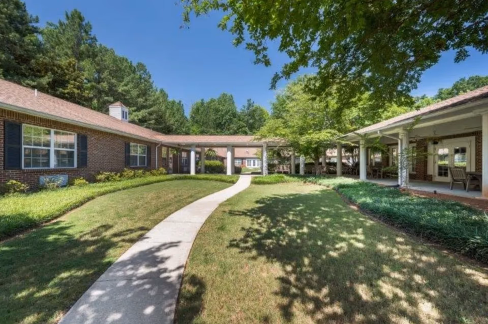 Outdoor view of Briar Glen Alzheimer's Special Care Center showing a curved concrete pathway through a grassy courtyard surrounded by single-story brick buildings with large windows and covered walkways. Trees provide shade and greenery around the area under a clear blue sky.