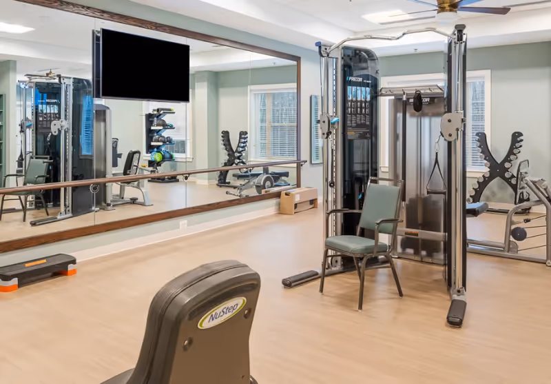 Well-equipped fitness room with weight machines, a NuStep exercise machine, dumbbells, a mirrored wall and a wall-mounted TV.