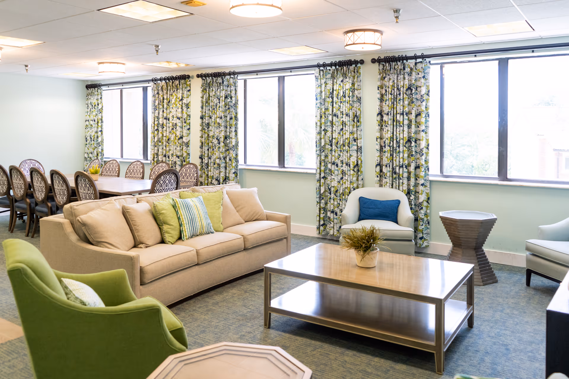 A bright and spacious living room area with a beige sofa adorned with green and striped pillows, a green armchair, two white armchairs with blue pillows, a wooden coffee table with a small plant, and a dining table with eight chairs in the background. The room has large windows with floral curtains allowing natural light to enter.