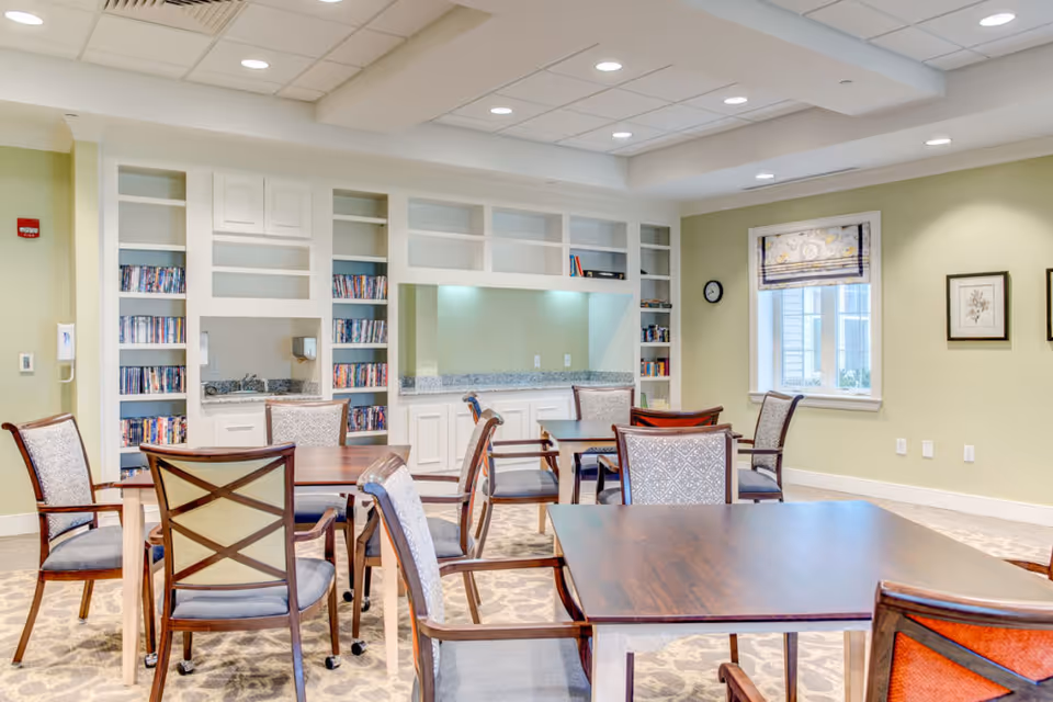 A bright and spacious common area with several wooden tables and cushioned chairs arranged around them. The room features built-in white shelves filled with DVDs and books, a countertop with a sink, and a window with a patterned Roman shade. The walls are painted light green, and the ceiling has recessed lighting.
