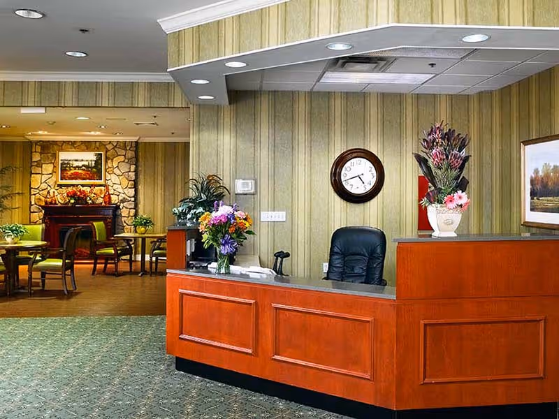 Reception area with a wooden front desk decorated with flower arrangements, a black office chair behind the desk, a wall clock showing 3:40, and a seating area with tables and chairs in the background near a stone fireplace with a framed painting above it.