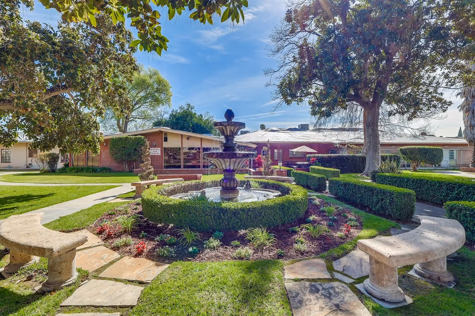 A sunny outdoor garden area at Colonial Gardens Nursing Home featuring a central tiered water fountain surrounded by neatly trimmed hedges and flower beds. Stone benches are placed around the fountain on a stone-paved path. Trees and well-maintained grass surround the garden, with a single-story brick building in the background under a clear blue sky.