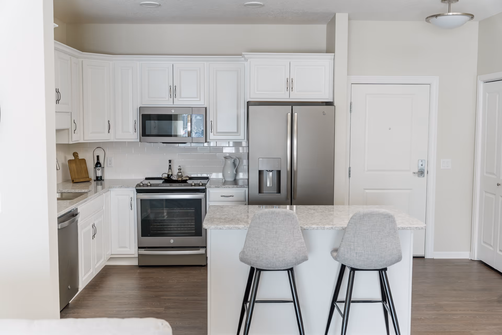 Modern kitchen with white cabinetry, stainless steel appliances including a refrigerator, oven, microwave, and dishwasher. There is a kitchen island with a marble countertop and two gray upholstered bar stools. The floor is wooden, and there is a white door and closet door in the background.