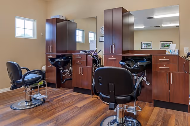 Interior view of a salon area with two black salon chairs in front of wooden cabinetry and sinks for hair washing. The room has wood flooring, beige walls, a large mirror, and two windows letting in natural light.