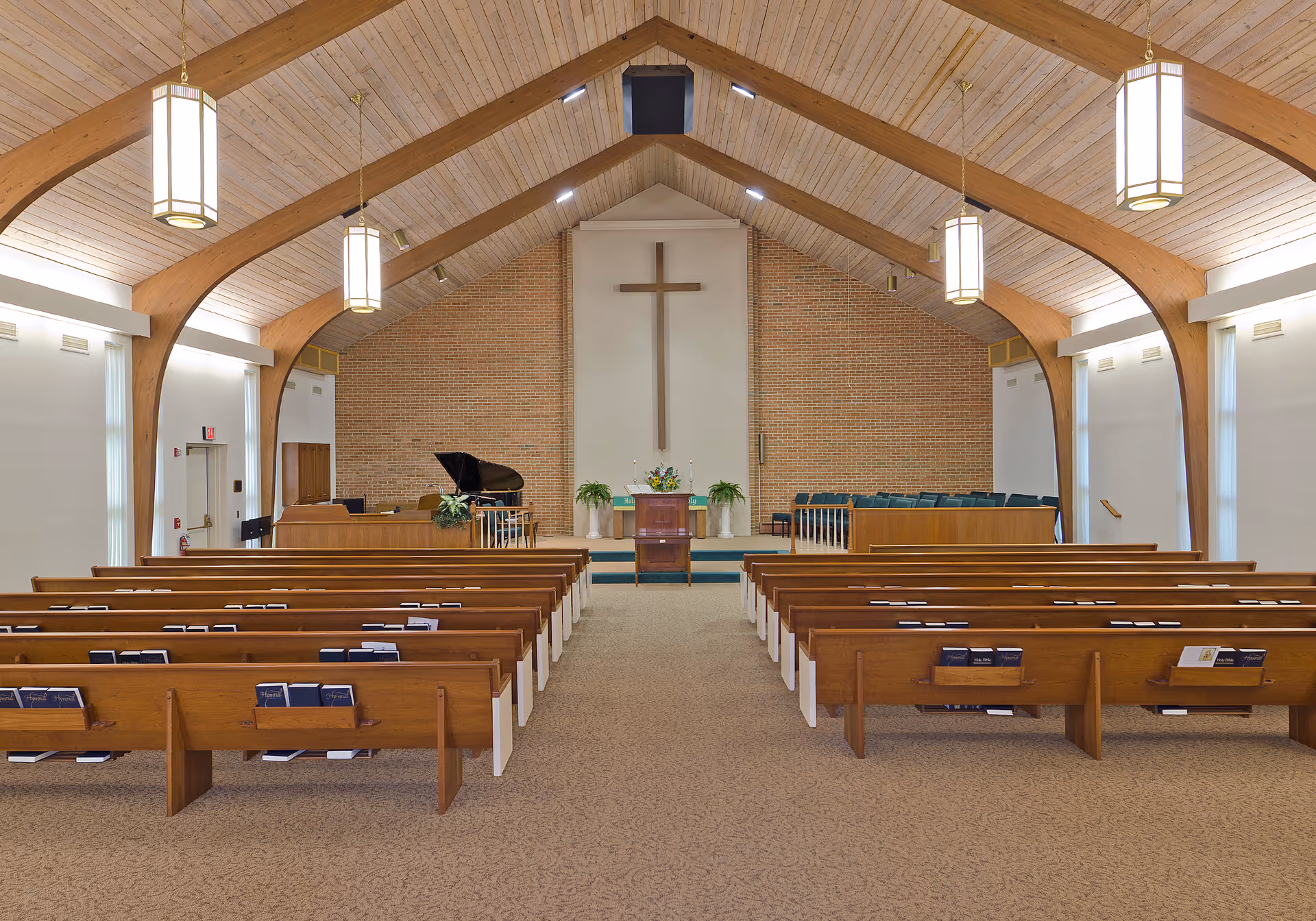 Interior of a chapel with wooden pews facing a raised altar area with a cross, piano, and vaulted wooden ceiling.