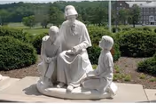 A white statue depicting an elderly person seated and reading a book to two children, one standing and one kneeling, set outdoors with greenery and buildings in the background.