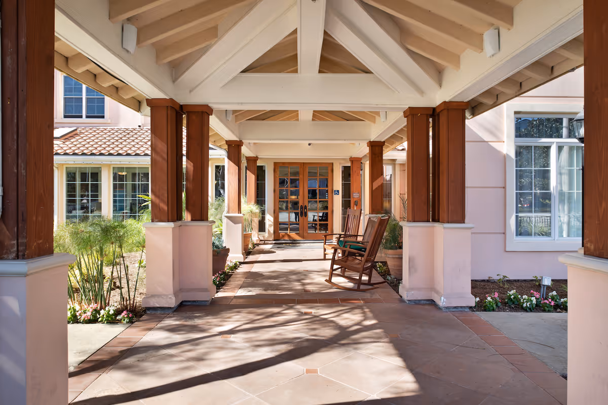 Covered entrance walkway with wooden columns, rocking chairs, and potted plants leading to glass double doors.