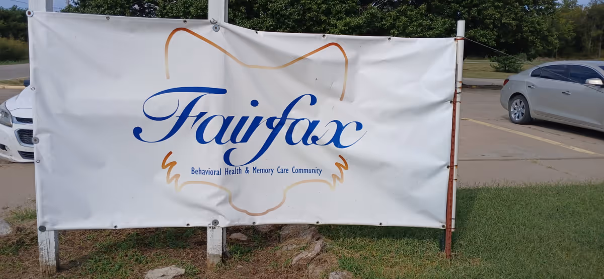 Outdoor banner reading "Fairfax Behavioral Health & Memory Care Community" mounted near a parking lot.