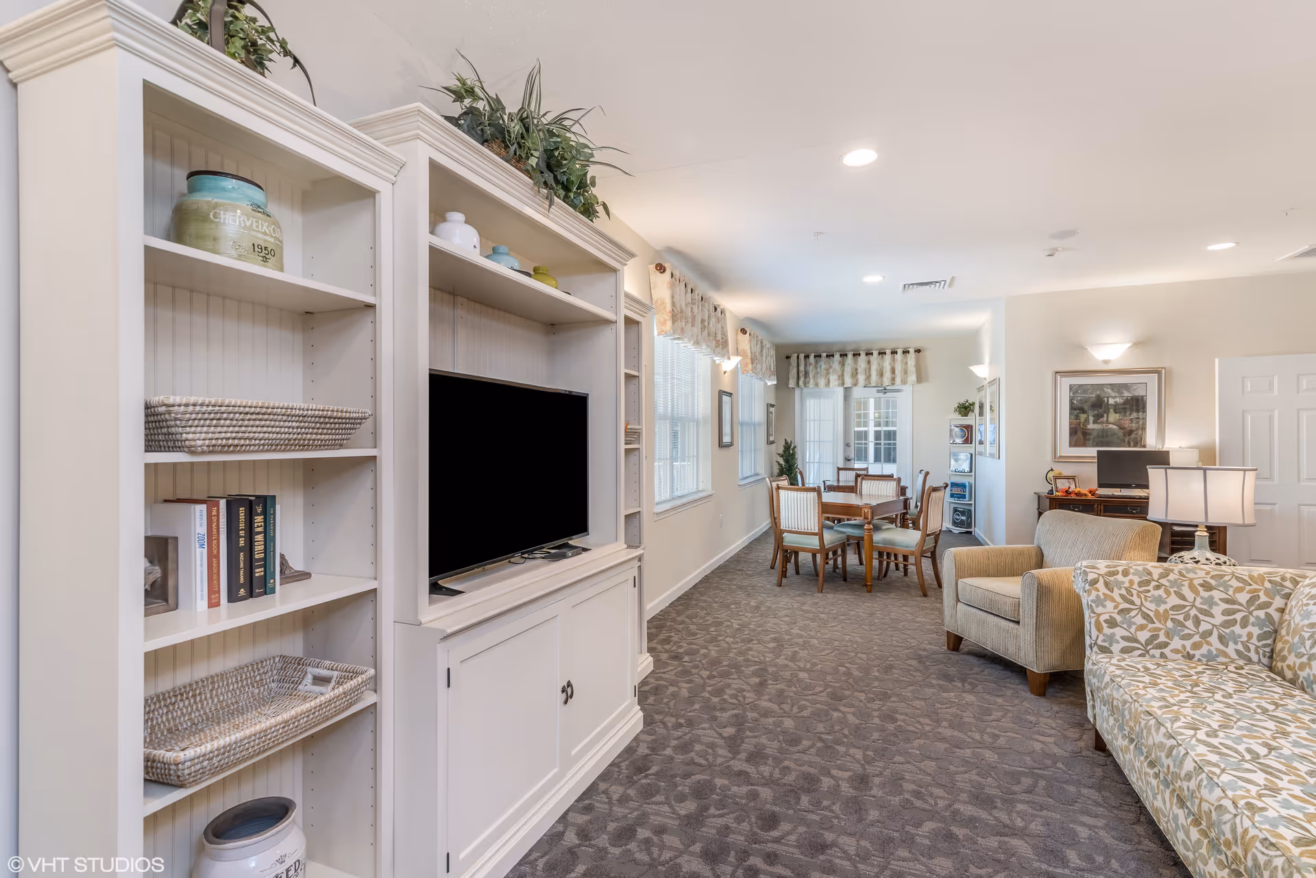A bright and spacious senior living common area featuring a white entertainment center with a flat-screen TV, shelves with decorative items and books, a patterned carpet, a floral-patterned sofa, an armchair, a table lamp, and a dining area with wooden chairs and tables near large windows with valances.
