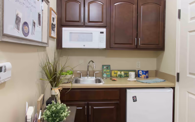 Small kitchenette area with dark wood cabinets, a white microwave mounted above a stainless steel sink, a beige countertop with decorative items including a small plant, a mug, and a box of crackers. There is a small white refrigerator below the counter and a whiteboard on the left wall.