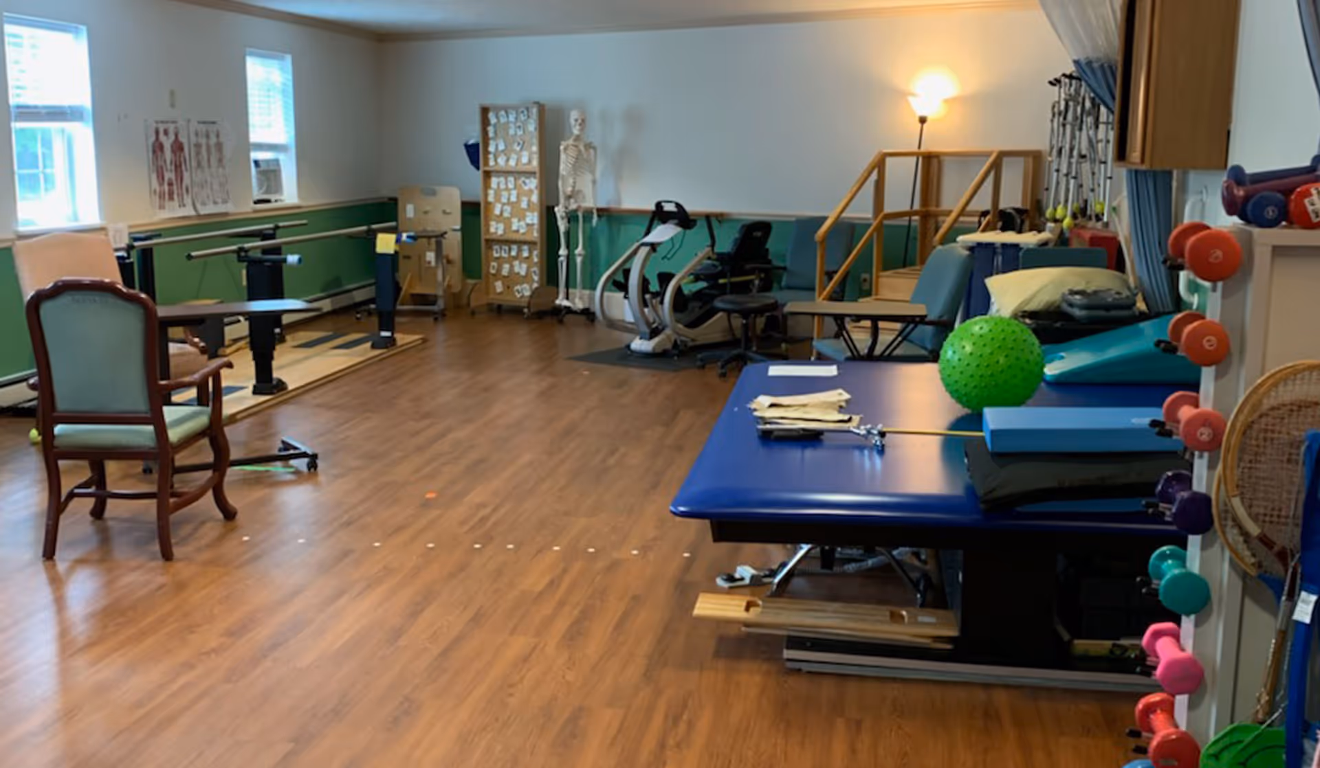 A physical therapy or rehabilitation room with wooden flooring, exercise equipment including parallel bars, stationary bikes, therapy tables with cushions, a green exercise ball, and various small dumbbells. There is a skeleton model and anatomical charts on the walls, along with a chair and a lamp in the corner.