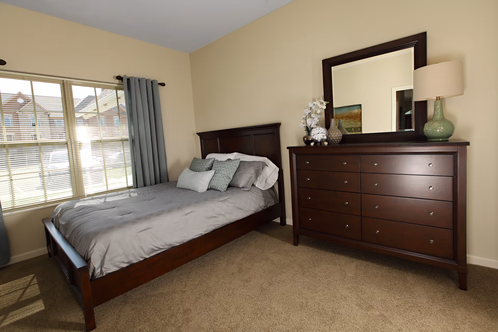 A bedroom with a wooden bed frame and gray bedding, several pillows, a large window with gray curtains, and a wooden dresser with a mirror, a lamp, a clock, and decorative items on top.